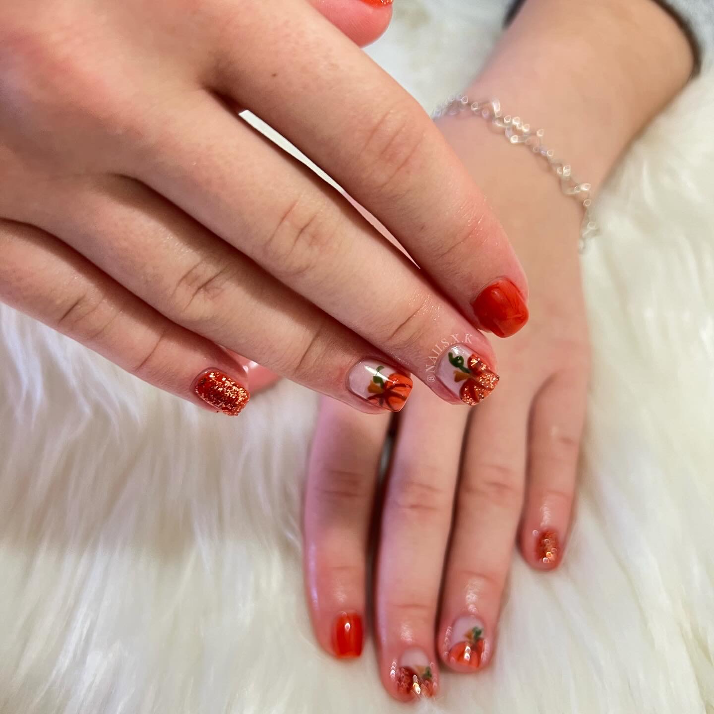 Gel manicure in a rust-orange color with hand-painted pumpkin designs and glitter accents. Nails by K
