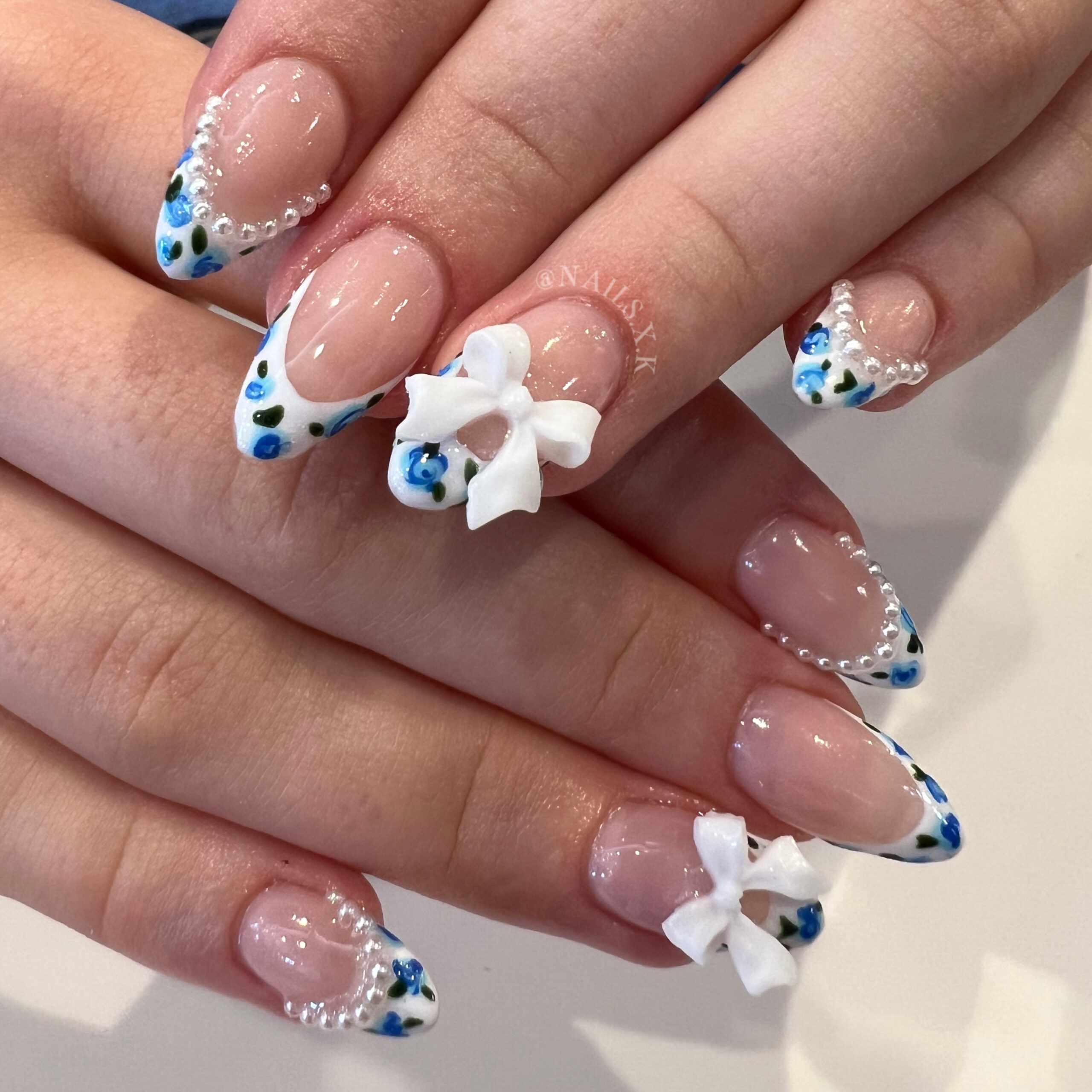 Acrylic nails with white french tips accented with hand painted blue rosettes, pearls and white bow charms. Nails by K